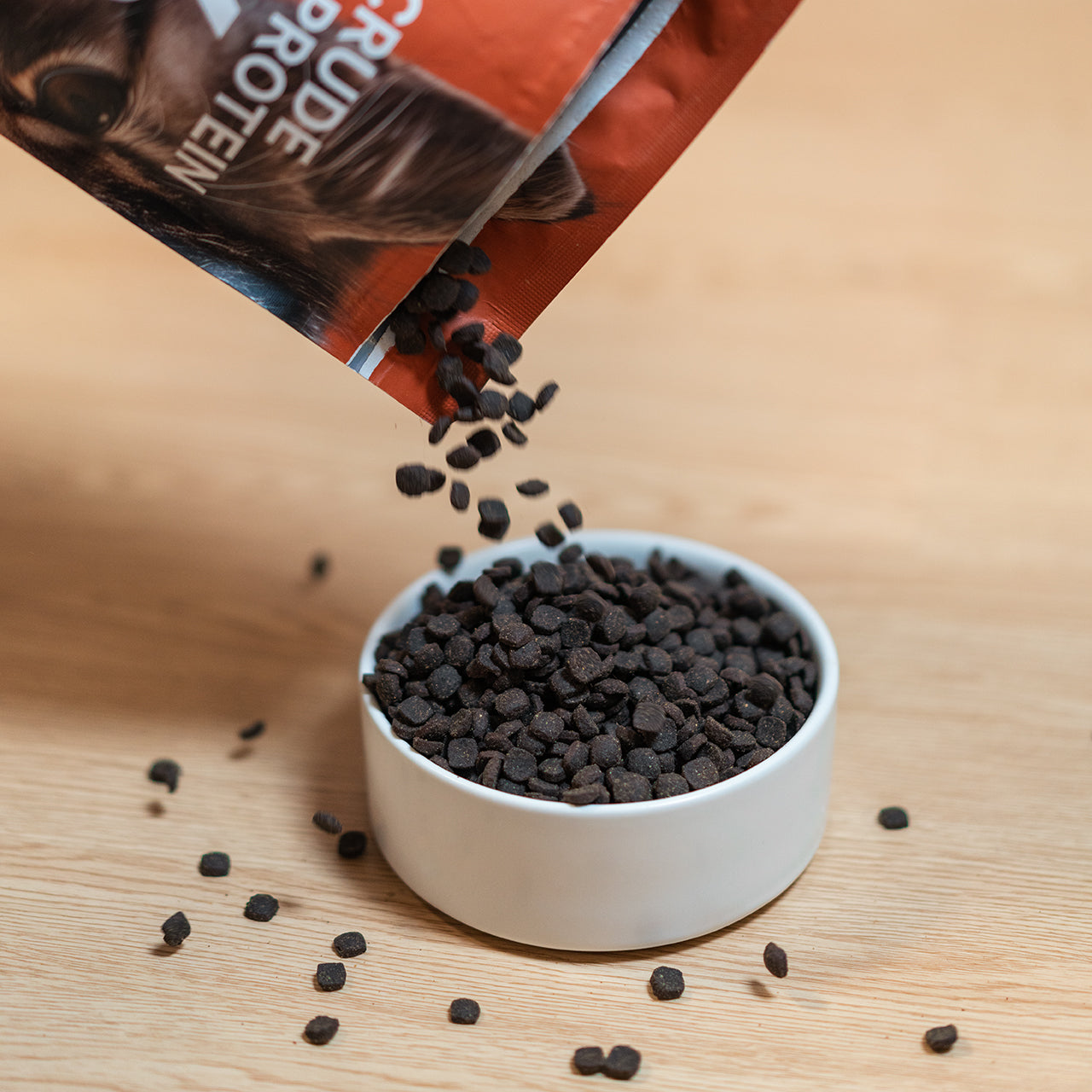 Pet food pouring into white bowl with grains scattered on wooden tabletop.